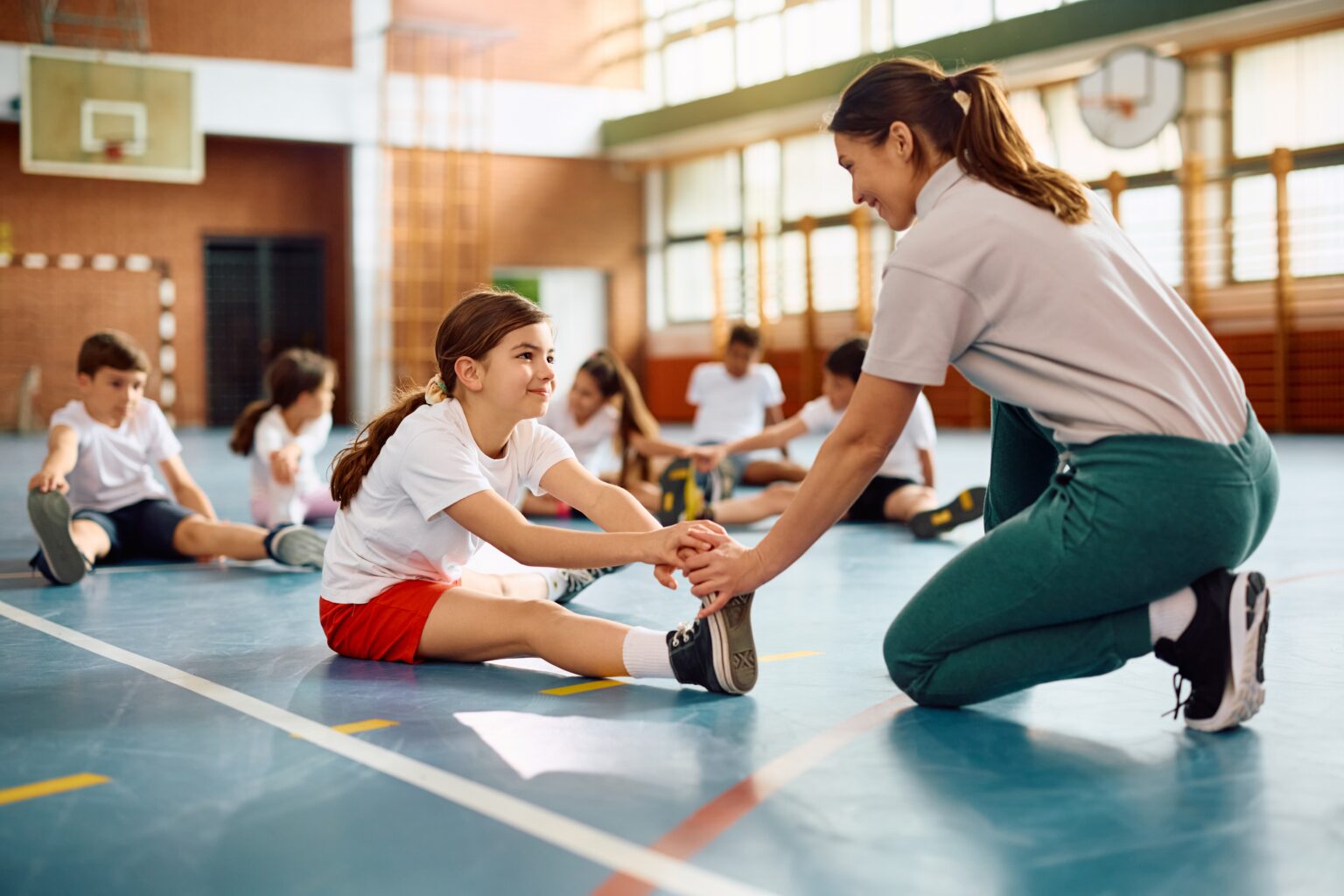 Happy,Schoolgirl,Stretching,Her,Leg,With,Teacher's,Assistance,On,Physical